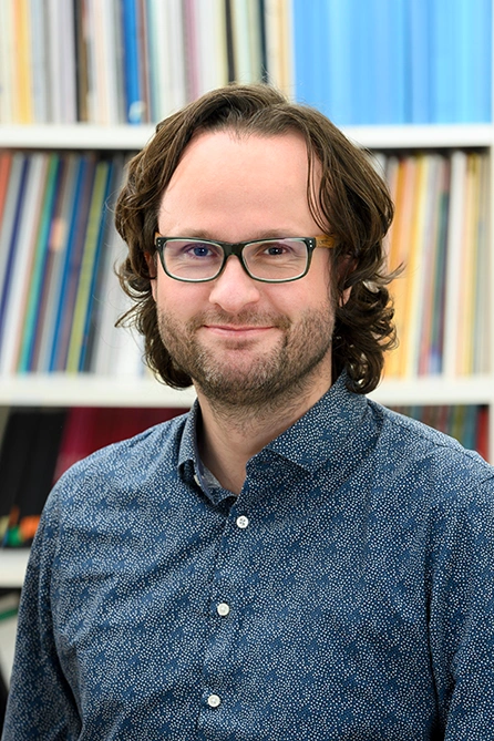 Man with medium-length curly hair and glasses, wearing a patterned shirt, standing in front of a bookshelf.