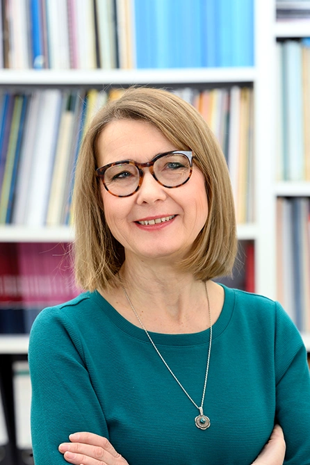 Woman with chin-length blonde hair and glasses, wearing a turquoise top, standing with arms crossed in front of a bookshelf.