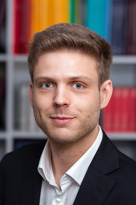 Portrait of a young man in a white shirt and dark jacket in front of a bookshelf with colorful spines.