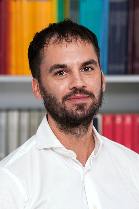 Portrait of a bearded man in a white shirt in front of a bookshelf with colorful spines.