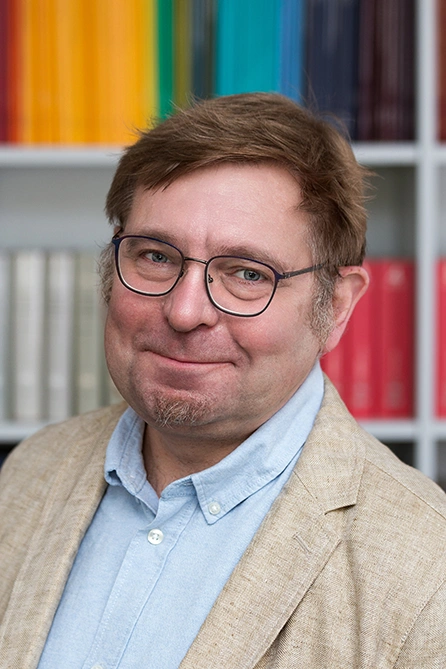 Mann mit kurzem braunem Haar und Brille, trägt ein beigefarbenes Sakko über einem hellblauen Hemd. Er steht vor einem Bücherregal mit farbigen Buchrücken und blickt freundlich in die Kamera.