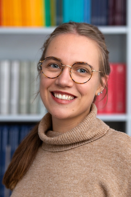 A young woman with glasses is smiling at the camera. She is wearing a beige turtleneck sweater and standing in front of a bookshelf filled with colorful book spines. The portrait appears friendly and professional.