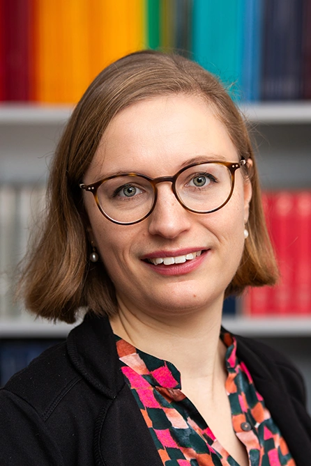 A woman wearing glasses is smiling at the camera. She is dressed in a colorful patterned blouse and a dark jacket. A bookshelf with color-coordinated spines is visible in the background.