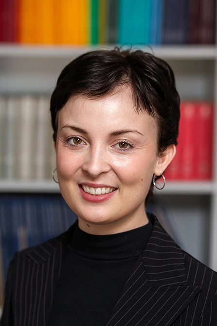 Woman with short dark hair and hoop earrings in front of a bookshelf, wearing a black turtleneck and dark pinstriped blazer.