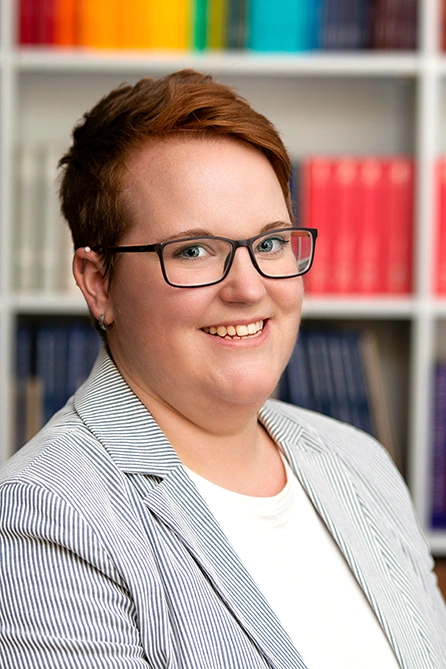 Frau mit kurzen rötlichWoman with short reddish-brown hair and glasses in front of a bookshelf, wearing a white top and a light pinstriped blazer.-braunen Haaren und Brille vor einem Bücherregal, trägt ein weißes Oberteil und einen hellen Blazer mit feinen Streifen.