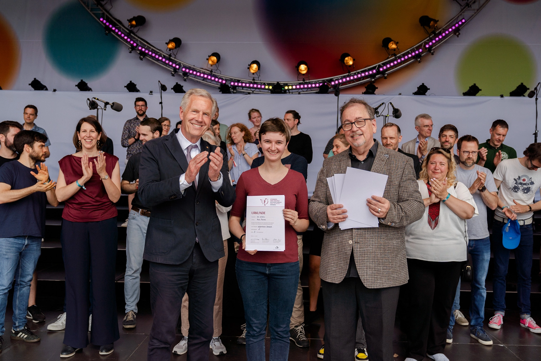 Eine Person erhält auf einer Bühne eine Urkunde, flankiert von zwei Männern, im Hintergrund applaudiert eine Gruppe. A person receives a certificate on stage, flanked by two men, with a group applauding in the background.