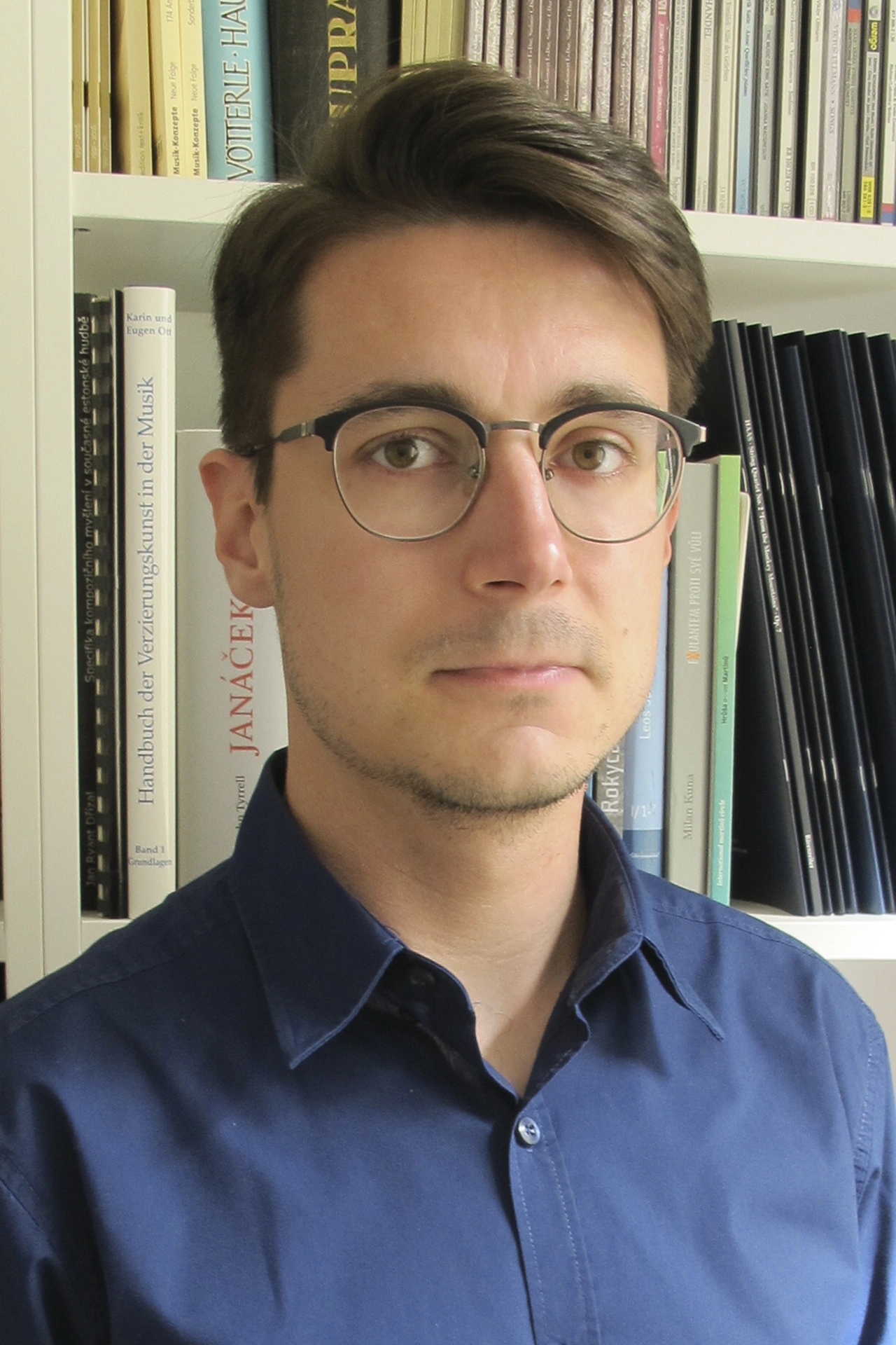 Young man with glasses and dark blue shirt stands in front of a bookshelf.