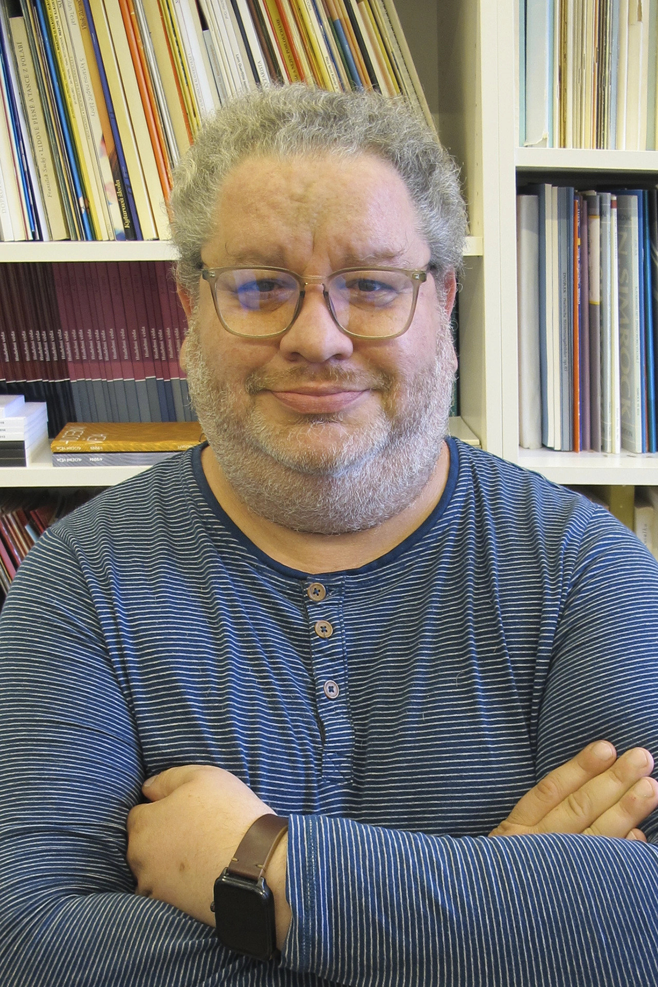 Man with glasses and striped shirt stands with crossed arms in front of a shelf with sheet music.